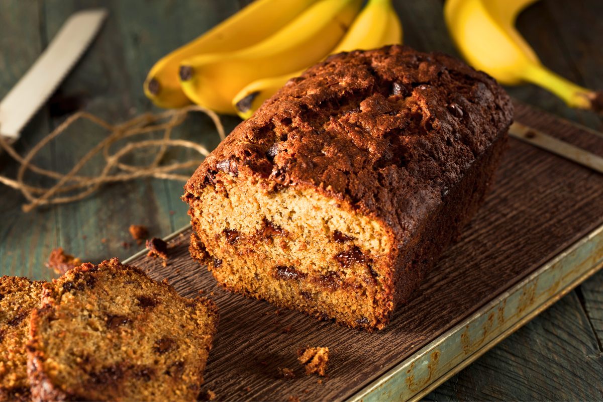 A loaf of chocolate chip banana bread with one slice cut up and some bananas in the background.