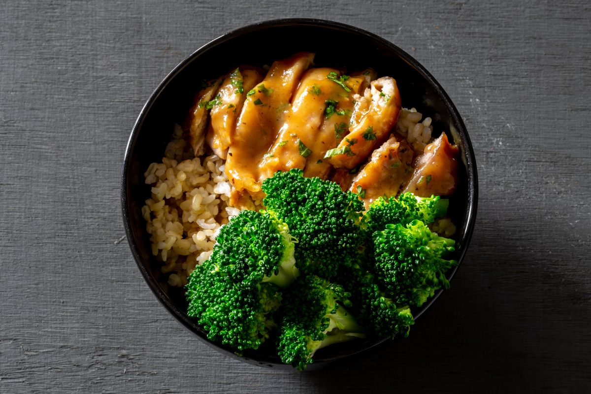 A bowl with honey mustard baked chicken breast, sliced, over brown rice accompanied with broccoli florest.