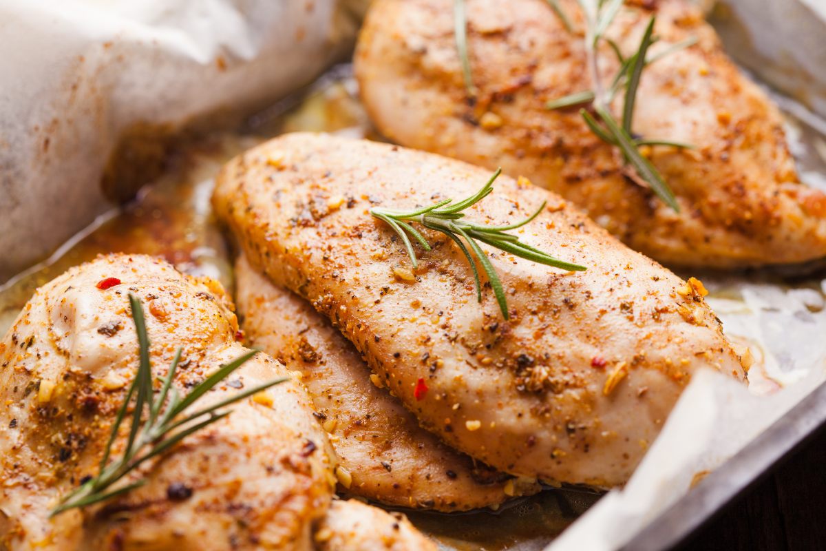 Baked chicken breasts with rosemary in an oven tray lined with paper.