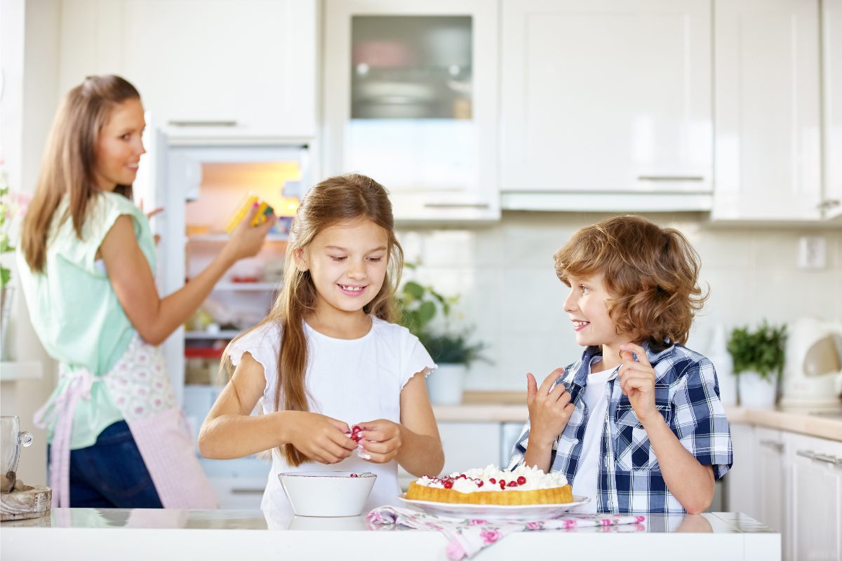 Two children decorating a cake with fruit and cream while the mother watches them.