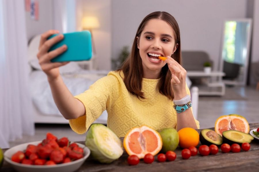 Inspired teen blogger chewing a carrot and using a phone to take the photograph.