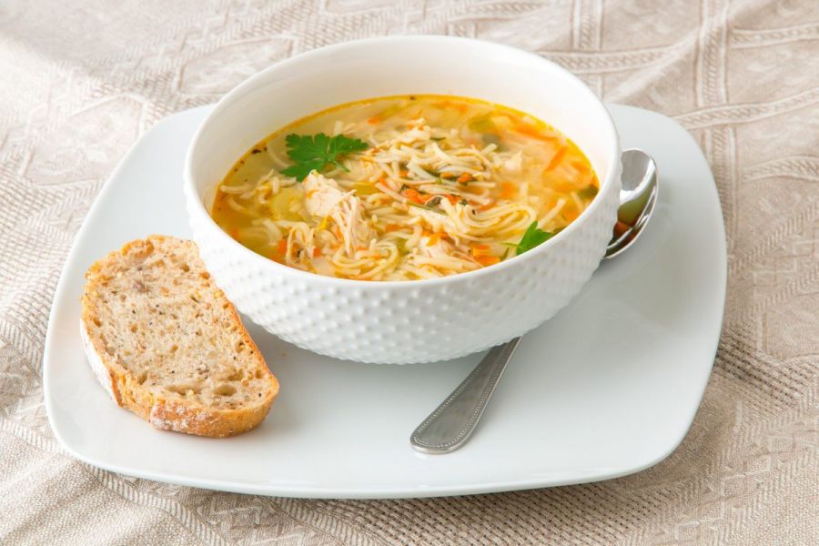 A bowl of chicken noodle soup on a plate with a slice of bread by the side.