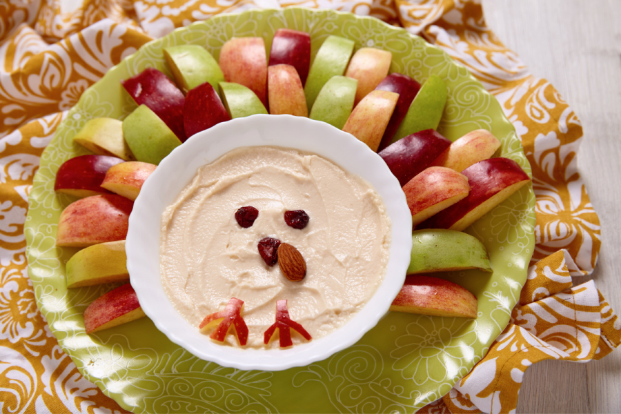 Caramel cream cheese dip in a bowl with dried cranberries as eyes and beak surrounded by apple wedges styled as the fan tail of a peacock.