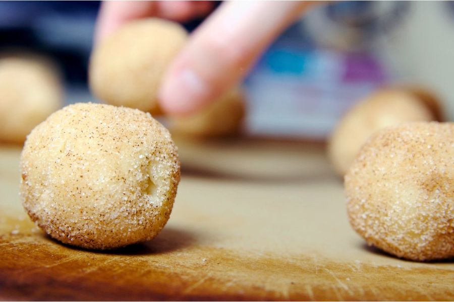 Close-up of snickerdoodle cookie dough balls rolled in cinnamon sugar, with a hand reaching for one in the background.